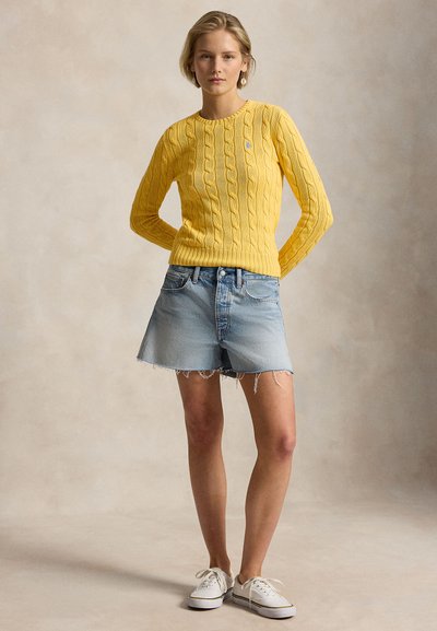 Γυναίκα όρθια με τα χέρια πίσω από την πλάτη, φοράει κίτρινο πουλόβερ με πλεκτό καλώδιο, ανοιχτόχρωμα τζιν σορτς, λευκά αθλητικά παπούτσια και σκουλαρίκια με μαργαριτάρια.