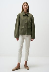 Giacca leggera verde oliva con colletto, maniche lunghe, due tasche frontali e taglio dritto; abbinata a pantaloni slim bianchi e scarpe beige.