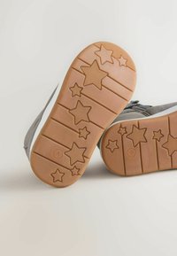 Scarpe in suede grigio con suole in gomma, caratterizzate da un motivo a stelle e un accento bianco. La suola è marrone con linee texture e forme di stelle.