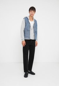 Vesta din denim peste un hanorac gri, asortată cu un tricou alb și pantaloni negri. Modelul poartă pantofi negri, stând în fața unui fundal alb.
