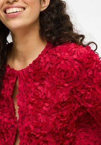 Veste texturée rouge à fleurs avec des pétales de tissu superposés, présentant un col rond et une ouverture en forme de clé à l'avant.
