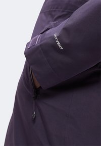 Gros plan d'une manche de veste violet foncé avec une sangle en velcro et le texte "DRY VENT" au-dessus d'une fermeture éclair partiellement visible.
