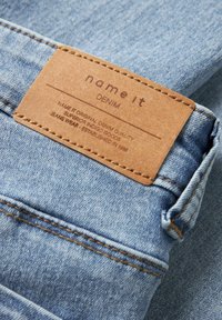 Džíny z denimu v světlém modrém odstínu, s hnědou koženou štítkem nesoucím nápis „name it“ a pokyny pro péči. Jsou viditelné přešívané švy.