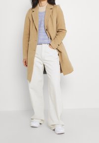 Manteau en laine beige au design ajusté, superposé sur un pull léger bleu, associé à un jean large blanc cassé et des baskets blanches.