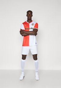 Rot und weißes Sporttrikot mit kurzen Ärmeln, versehen mit einem Logo und dem Aufdruck "MediaMarkt". Kombiniert mit weißen Shorts und Socken sowie weißen Fußbällen.
