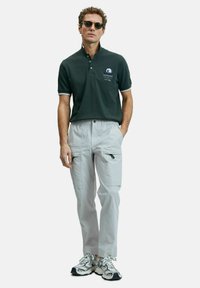 Donkergroene polo met witte accenten en logo, gecombineerd met lichtgrijze cargobroek met zijzakken en ritsen, afgewerkt met sneakers.