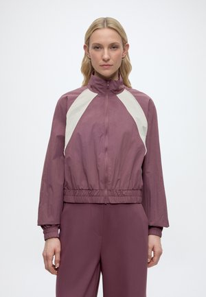 Donna con capelli biondi lisci che indossa una giacca mauve con zip e pannelli bianchi a chevron e pantaloni mauve abbinati, in piedi su uno sfondo bianco.