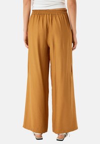 Wijdflatige broek van een goudbruine stof. Kenmerkt zich door een elastische tailleband en zijsplitten. Soepele textuur met een ontspannen silhouet.