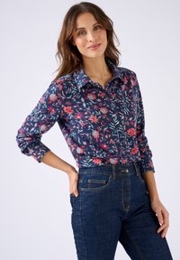 Donna che indossa una camicia a maniche lunghe blu con fantasia floreale, infilata in un paio di jeans scuri a vita alta, in piedi con una mano sul fianco contro uno sfondo neutro.