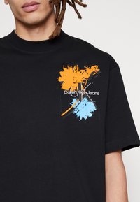 Black cotton t-shirt featuring a colorful graphic design with orange and blue paint splatters, and "Calvin Klein Jeans" logo in white text.