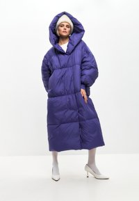 Ljubičasti kaput s podstavom i velikom kapuljačom, s naglašenim strukom i velikim džepovima, nošen s bijelim petama i teksturiranim čarapama.