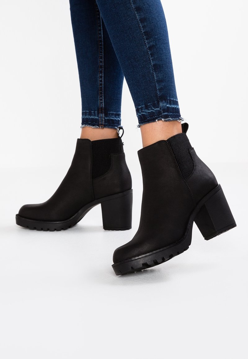 Personne portant des bottines en cuir noir à talons blocs et panneaux latéraux élastiques associées à un jean skinny bleu foncé.