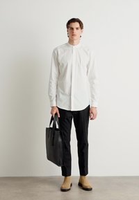 Bela dolgimi rokavi, slim-fit črnimi hlačami, svetlimi gležnjarji, drži črno usnjeno torbo, v ozadju svetla podlaga.