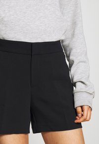 Černé, tailored šortky s hladkou texturou, mají plochý pas a jemný přední šev. Oblečení je doplněno šedou mikinou.
