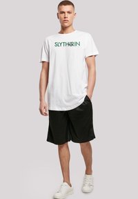 Hvid bomulds T-shirt med grøn "SLYTH3RIN" grafisk tekst, parret med sorte mesh shorts og hvide sneakers. Enkel design, afslappet pasform.