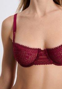 Bordowy stanik koronkowy z kwiatowymi wzorami, falbaniastymi brzegami, regulowanymi ramiączkami oraz zapięciem na haftkę z tyłu.