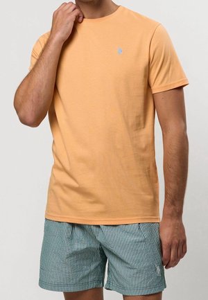 Mand iført ferskenfarvet t-shirt med korte ærmer og grønne og hvide stribede shorts, hånd løftet nær ansigtet, mod ensfarvet baggrund.