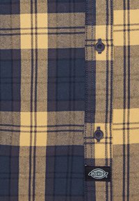 Tissu de chemise à carreaux jaune et bleu foncé avec deux boutons bleu marine et une petite étiquette noire de la marque Dickies cousue près de la couture.