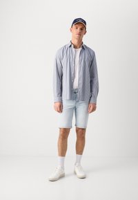 Chemise rayée bleu et blanc sur un T-shirt blanc, associée à un short en denim léger et des baskets blanches. Une casquette bleue complète le look.