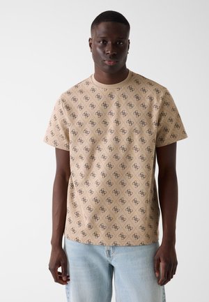 Junger Mann, der nach vorne schaut, trägt ein beigefarbenes Kurzarmhemd mit dunklen, gemusterten Logos und hellblaue Jeans vor einem schlichten weißen Hintergrund.