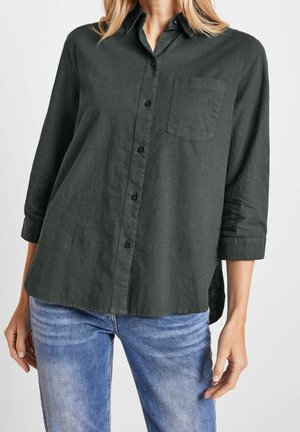 Femme portant une chemise gris foncé boutonnée avec les manches retroussées et un jean bleu clair, debout devant un fond uni.