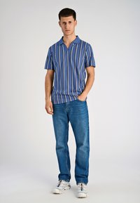 Koszula z krótkim rękawem w niebieskie i białe paski, zapinana na dekolt w serek, w połączeniu z niebieskimi dżinsami i białymi sportowymi butami.