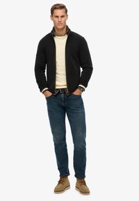 Zwarte zip-up jas, crème trui, blauwe spijkerbroek, tan laarzen. Jas heeft elastische manchetten en twee voorzakken. Staande kraag met groene accenten.