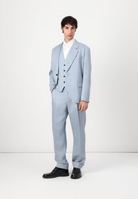 Traje azul claro de tres piezas con chaqueta de solapa muesca, chaleco abotonado y pantalones de pierna ancha, complementado con zapatos de vestir negros.