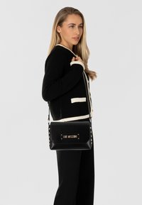 Borsa a mano in pelle nera con una placca logo dorata e dettagli borchiati, caratterizzata da una forma rettangolare elegante e una tracolla removibile.