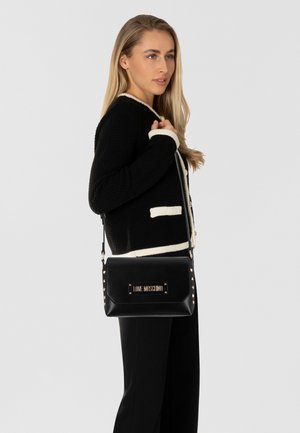 Borsa a mano in pelle nera con una placca logo dorata e dettagli borchiati, caratterizzata da una forma rettangolare elegante e una tracolla removibile.