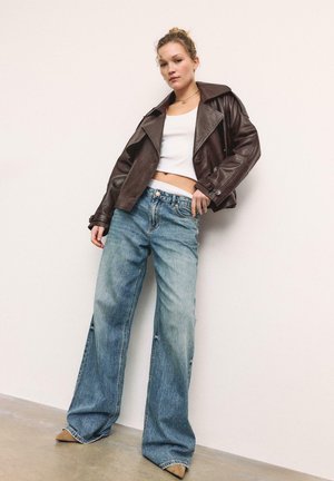 Giovane donna appoggiata al muro che indossa una giacca di pelle marrone scuro, un top bianco corto, jeans a gamba larga blu e scarpe beige.