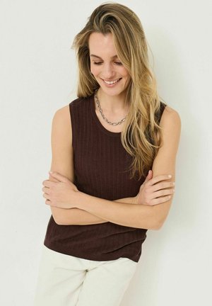 Femme souriante aux longs cheveux blonds, portant un haut sans manches en maille marron, un collier en chaîne argentée et un pantalon blanc, debout les bras croisés.