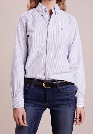 Femme portant une chemise boutonnée à rayures bleu clair et blanches, rentrée dans un jean bleu foncé avec une ceinture noire à boucle dorée.