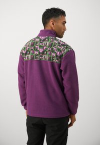 Lilla fleece-pullover med høy krage; mønstret grønn og rosa panel over skuldrene; avslappet passform med lange ermer.