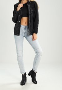 Chaqueta de mezclilla negra con detalles desgastados, combinada con un top negro corto y unos jeans skinny de color azul claro con parches en las rodillas. Botines negros.