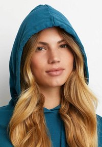 Felpa in cotone verde acqua con cappuccio regolabile, texture morbida e vestibilità rilassata. Il modello ha i capelli biondi lunghi e mossi con riflessi naturali.