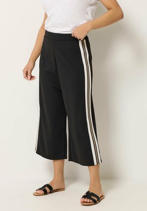 Sorte bredbenede cropped bukser med hvide sidelængder båret med hvid top og sorte flade sandaler på lys baggrund.