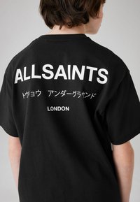 Černé bavlněné tričko s krátkým rukávem, na němž je na přední straně bílé text: "ALLSAINTS", japonské znaky a na zadní straně "LONDÝN".