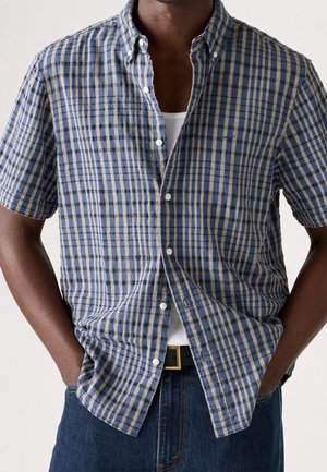 Uomo con camicia a maniche corte a quadri blu, marrone e bianca sopra una canottiera bianca con le mani nelle tasche dei jeans.