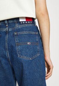 Džínové kalhoty modré barvy s kontrastním šitím. Obsahují logotyp v červené, bílé a modré na pásku a design zadní kapsy.