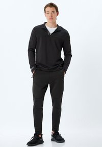 Sort zip-up langærmet top med en stribet detalje på kraven, parret med sorte tætsiddende bukser med knæpaneler. Sneakers fuldender outfittet.