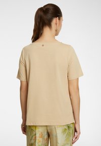 Camicia beige a maniche corte con scollatura a girocollo e un piccolo dettaglio decorativo sul retro. Tessuto morbido e vestibilità rilassata. Abbinata a pantaloni fantasia.