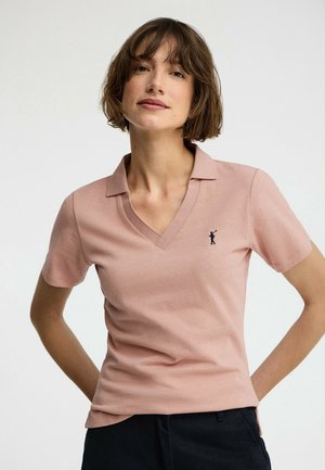 Kvinde med kort brunt hår iført en lyserød kortærmet polo med en lille sort golfspillerlogo på brystet, kombineret med mørke bukser.