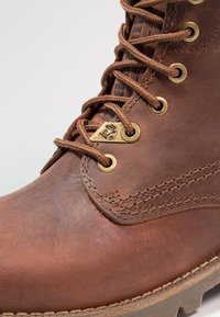 Botas de cuero marrón con ojales de latón, superficie texturizada y costura intrincada a lo largo del costado. Presenta cordones redondos para un ajuste seguro.