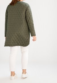 Quilted green jacket with a diagonal pattern, long sleeves, and a rounded hem, paired with white pants and beige sneakers.