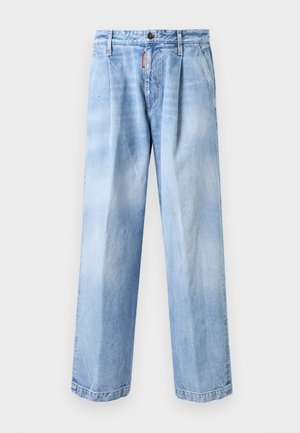 Muž nosiaci červené tričko s grafikou, voľné svetlomodré džínsy a biele tenisky stojí pri jednoduchom svetlom múre.