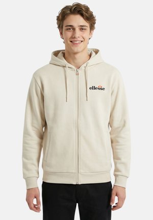 Jeune homme souriant, portant un sweat à capuche beige clair zippé avec le logo « ellesse » et un pantalon noir, debout sur fond blanc.