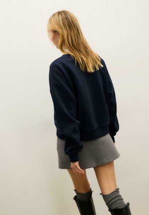 Mornarsko plavi kratki duks s spuštenim ramenima, u kombinaciji s sivom mini suknjom. Outfit uključuje slojevite čarape i crne čizme do koljena.