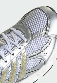 Baskets de sport argentées et blanches avec des surpiqûres métalliques, une tige en mesh respirant, des lacets blancs et trois bandes dorées texturées sur le côté.