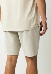 Pantaloni scurți gri deschis, dintr-un material texturat, cu o margine well finisată și buzunare laterale, purtați cu o cămașă bej.